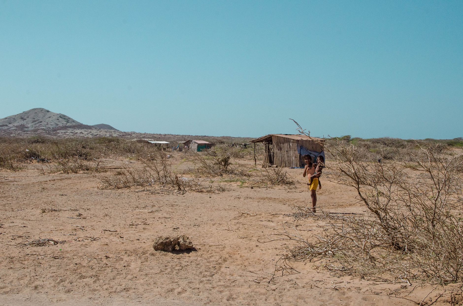 A child walks through an arid desert landscape near a minimalist wooden hut under a clear blue sky.