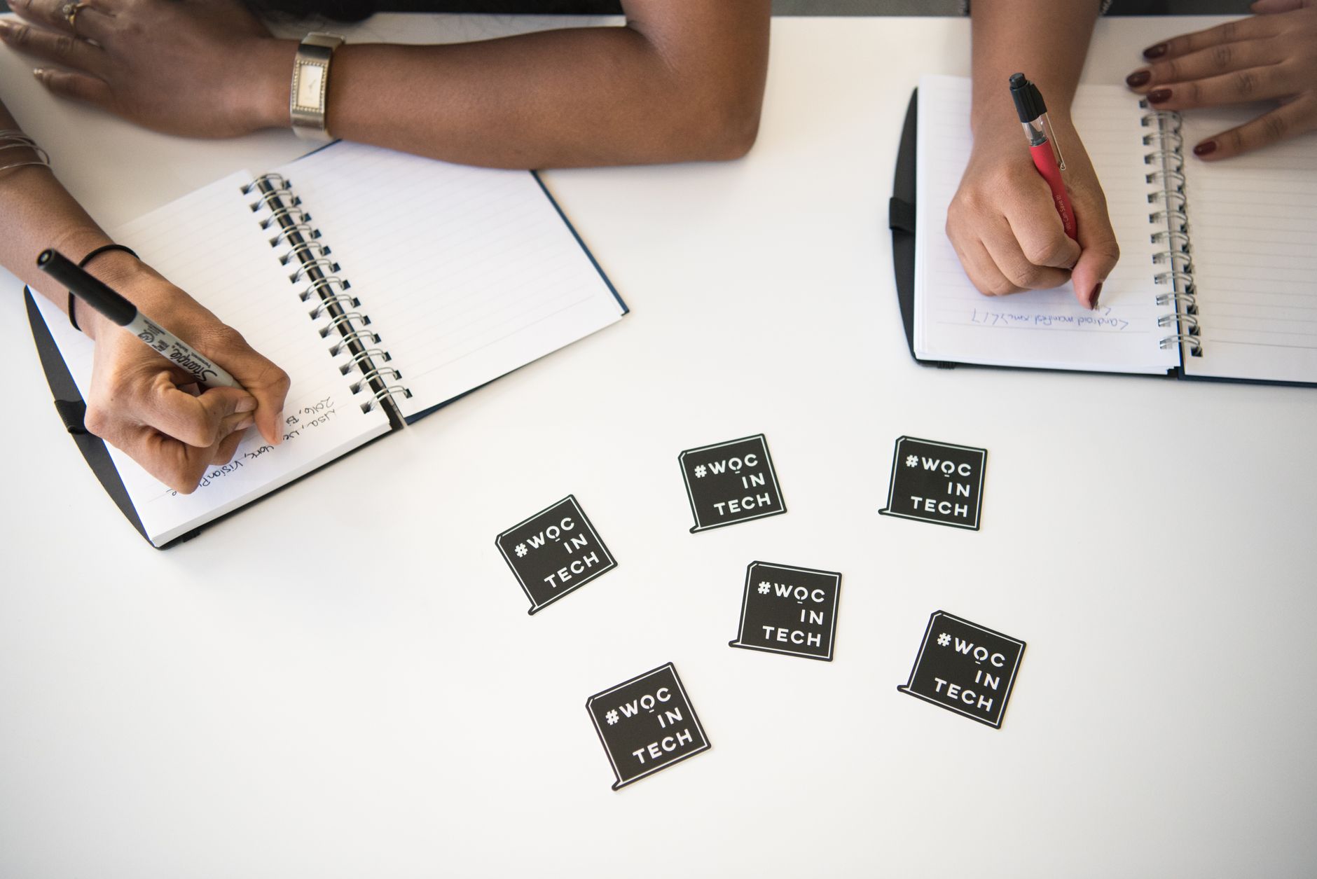 Diverse women collaborating in a tech meeting, writing notes in notebooks on a white desk.