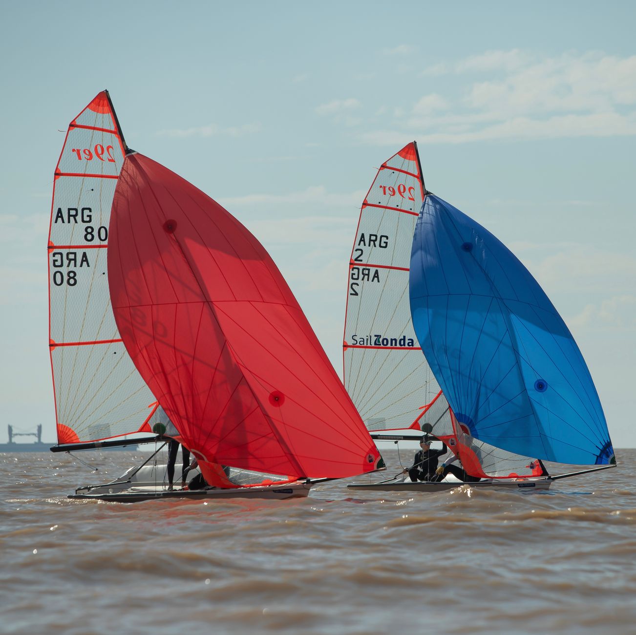 Two sailboats compete with red and blue sails on open water during the day.
