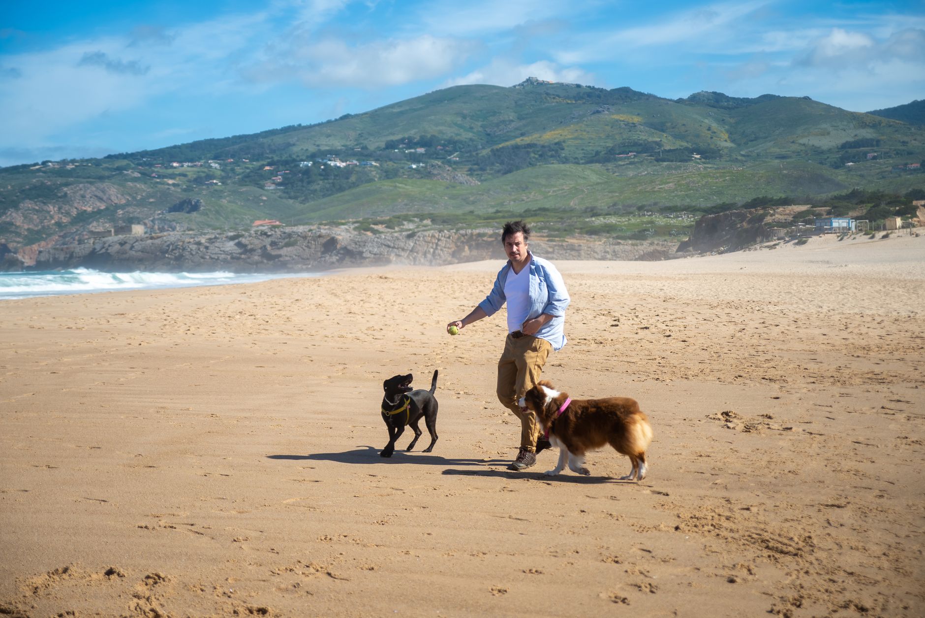 A man enjoys a sunny day playing fetch with two dogs on a picturesque beach in Portugal.