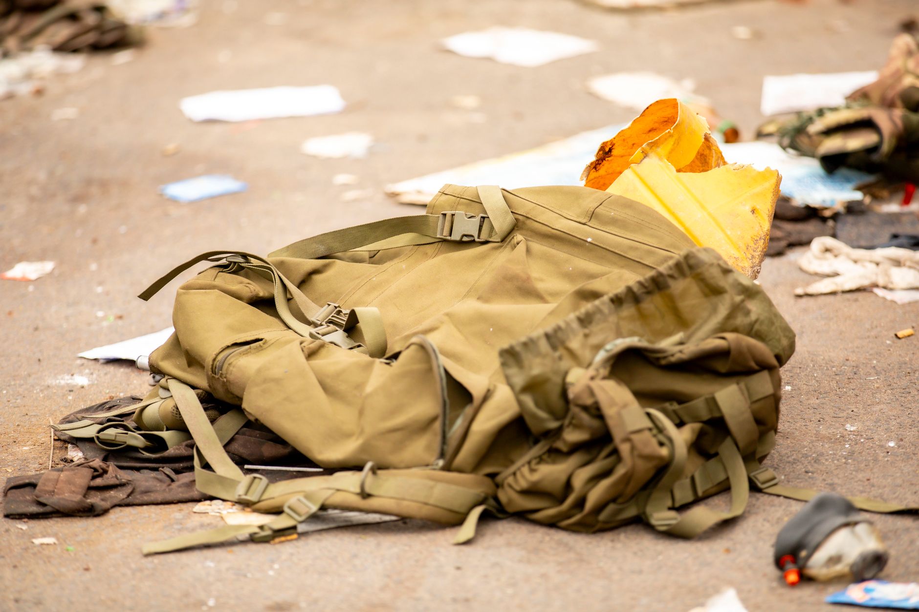 An abandoned military backpack lies on a trash-strewn street in an urban setting.