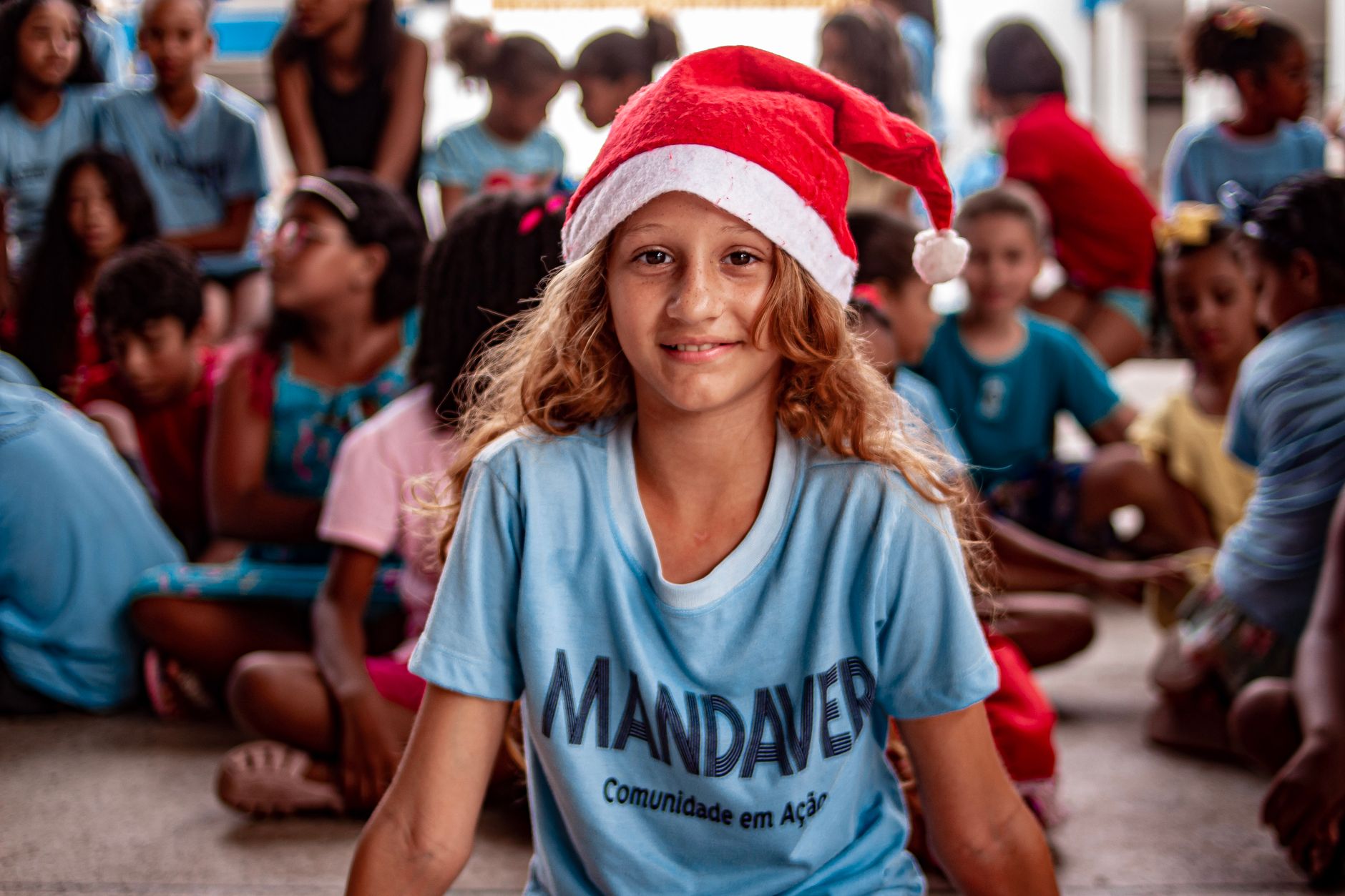 A smiling girl wearing a Santa hat, surrounded by a group of children, at an indoor holiday event.