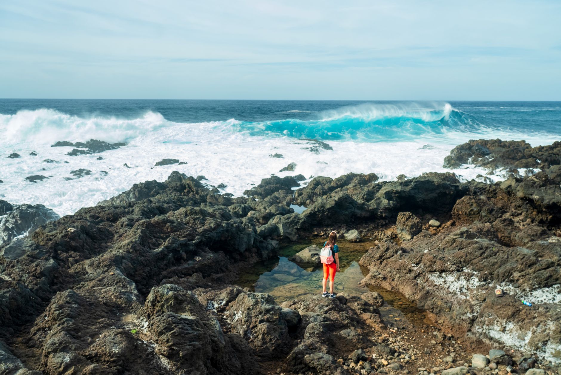 A woman stands on rocky terrain by the sea in El Sauzal, Spain, observing crashing waves under a clear sky.