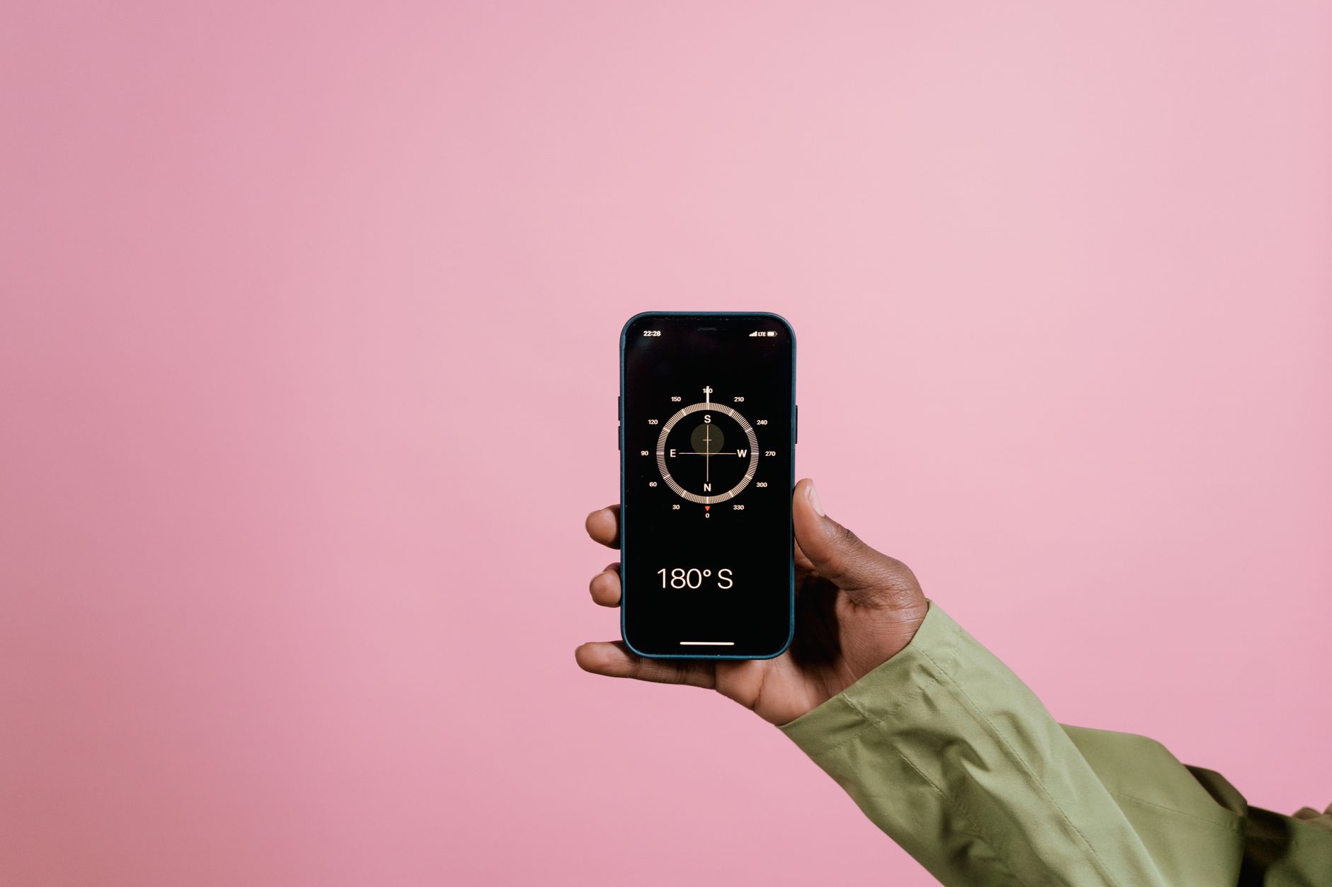 Close-up of a hand holding a smartphone displaying a compass app against a pink background.