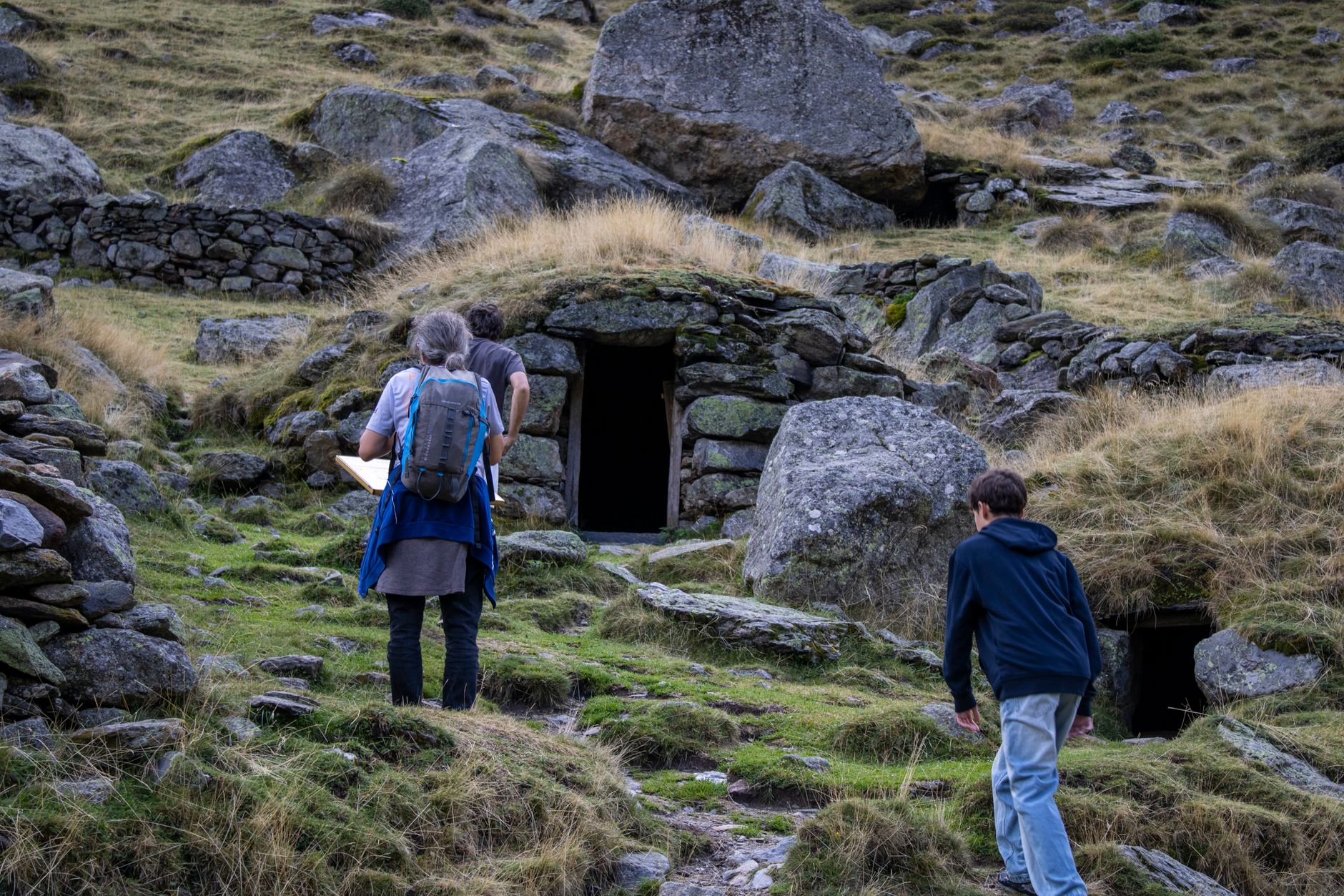 Two people exploring ancient stone huts built into rocky hills under a cloudy sky.