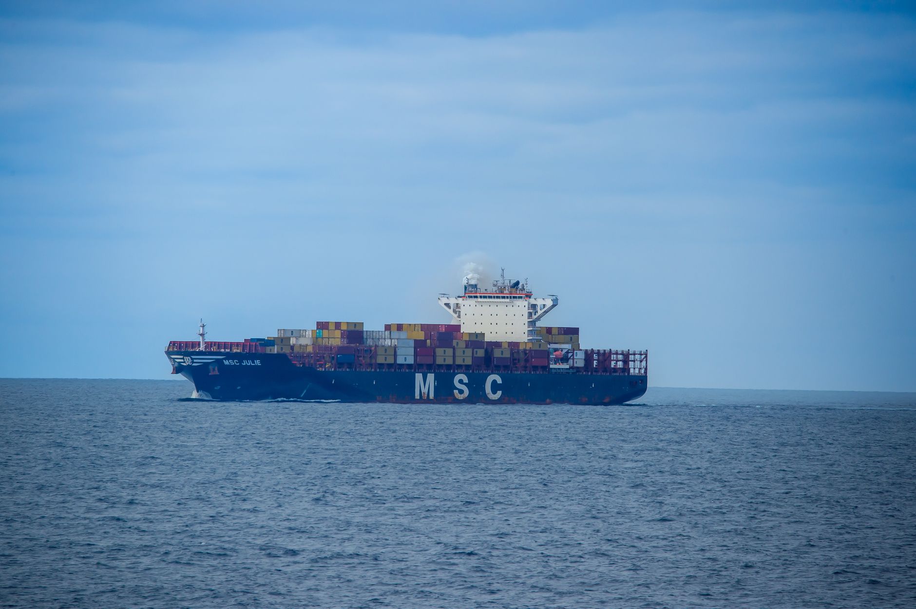 A large cargo ship sailing on a calm sea under a clear blue sky, MSC containers visible.