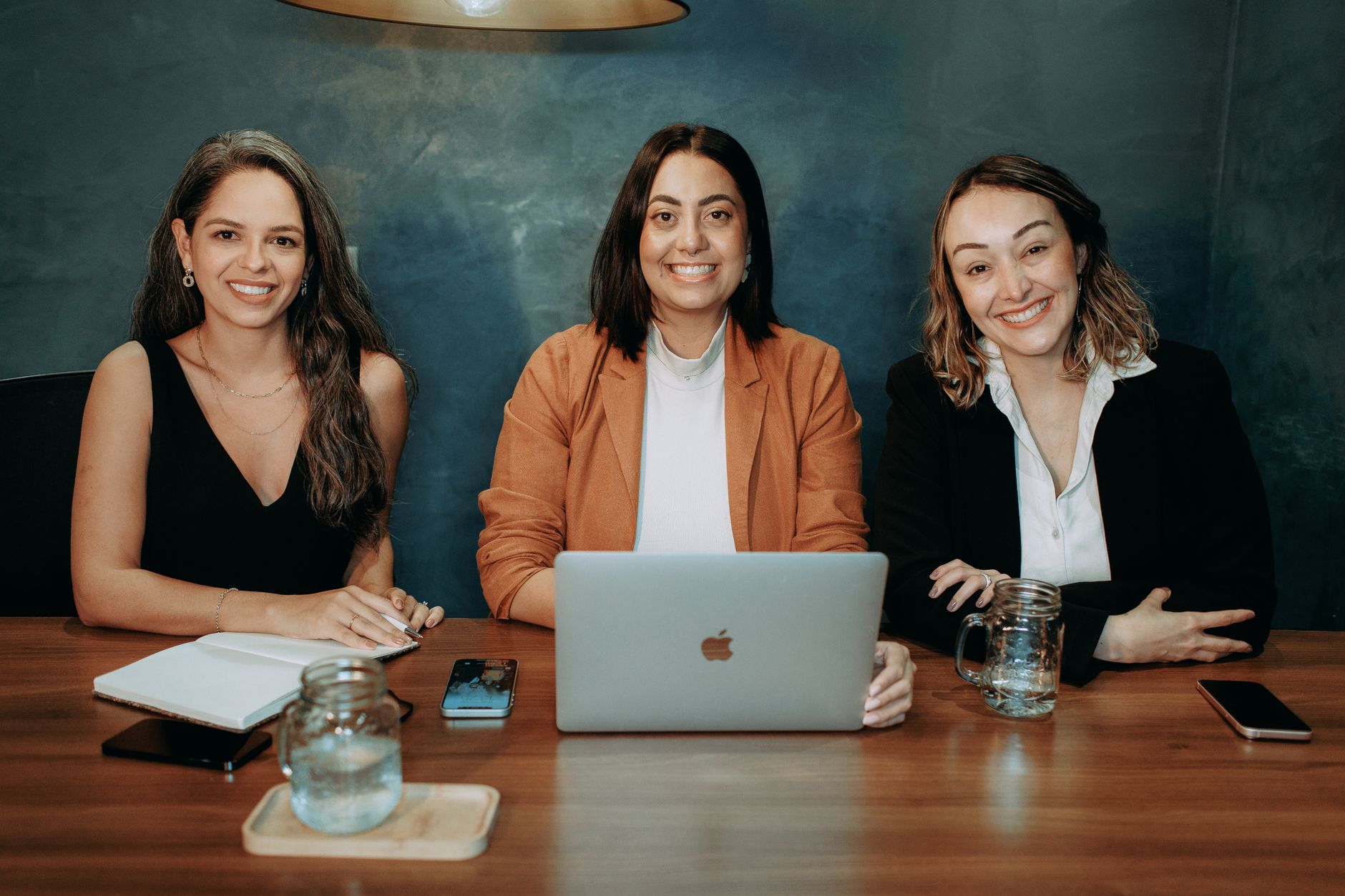 Three professional women sitting at a wooden desk in an office with laptops and notebooks.