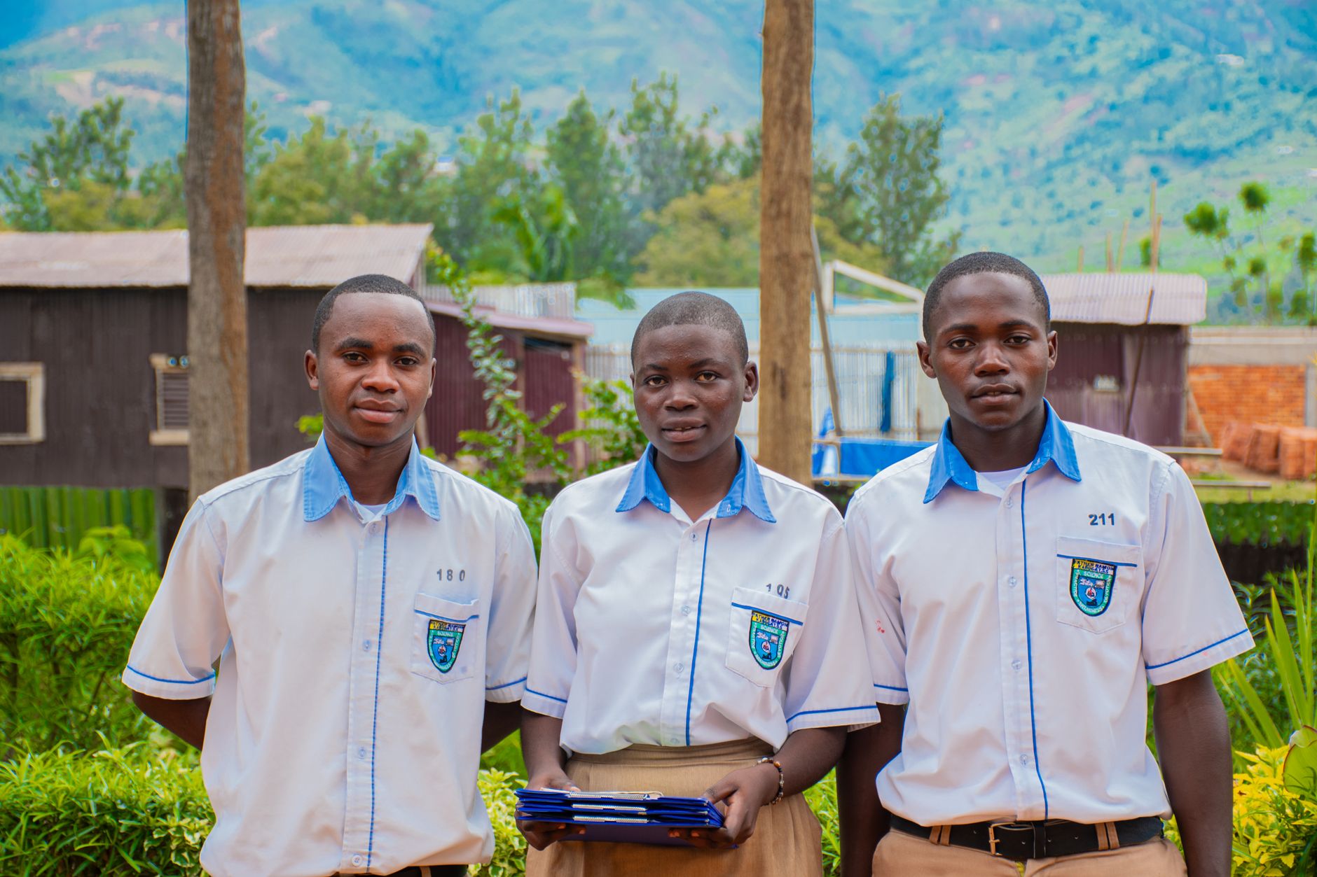 Three students in uniforms standing outdoors with a scenic mountain background.